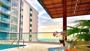 Uma área na piscina com vista para um edifício moderno, um espaço de estar coberto com uma mesa e um vaso de planta.