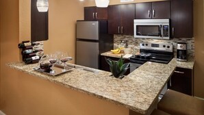 Eine Küche mit dunklen Holzschränken, einem Edelstahlkühlschrank, einer Mikrowelle, einem Backofen und einer Granit Arbeitsplatte. Auf der Arbeitsplatte stehen Weinflaschen und Gläser.