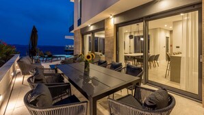 An outdoor dining area with a table and chairs, a view of the sea, and a glass door leading to an interior space. - Beachfront Villa Aurelia with heated Pool, Sauna & Fitness (Pijavičino (Pelješac))
