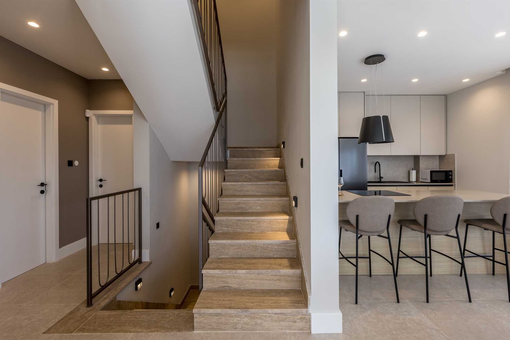A modern kitchen with a central island, bar stools, and a staircase leading to another level.