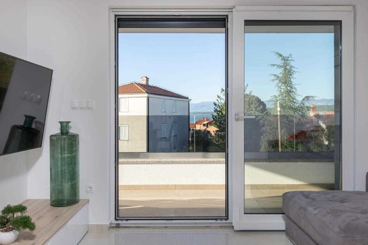 A room with a sliding glass door leading to a balcony, a small table with a green vase and a bonsai tree, and a bed with a grey headboard.