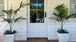 A white door with a glass panel, flanked by two potted plants, on a wooden porch with a white mat at the entrance. - Newly Renovated, Little Palm Cottage with dock slip, Elbow Cay  (Elbow Cay)