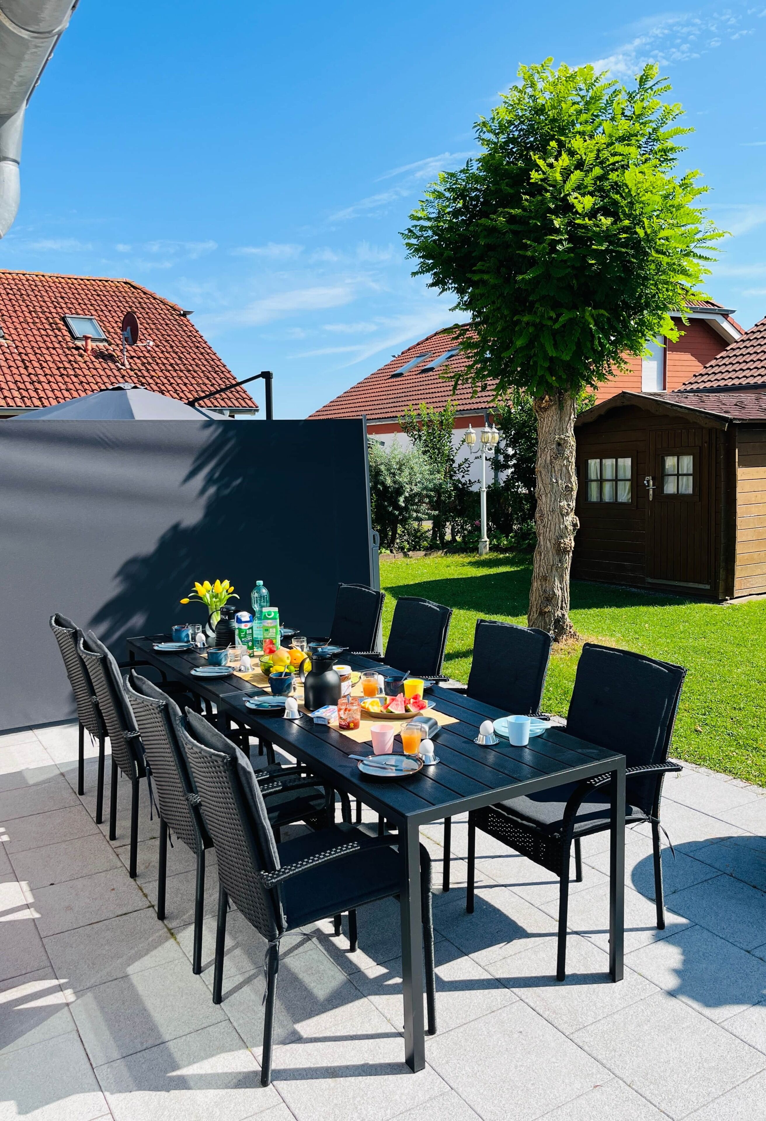 An outdoor dining area with a table set for six, surrounded by chairs, under a clear sky.