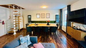 Una sala de estar moderna con una mesa de comedor de madera, sillas tapizadas en verde, un sofá gris con cojines estampados, una escalera de madera y un televisor de pantalla plana montado en la pared.