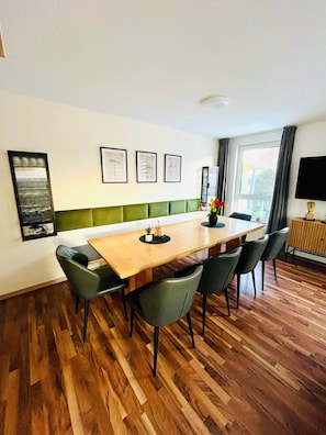 A dining area with a wooden table, green upholstered chairs, framed artwork on the wall, and a window with curtains.