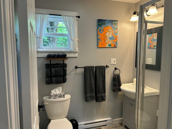 A bathroom with a toilet, sink, and two towels hanging on a rack. There is a window with curtains, a painting on the wall, and a mirror. - Quaco Cottages - The Waterloo
(West Quaco)