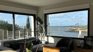 A modern living room with a large window overlooking a coastal landscape, a ceiling light fixture, and a wooden coffee table with a potted plant. - Perle des Abers, Profitez de Cette Charmante Maison de Vacances Avec vue mer (Saint-Pabu)