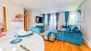 A modern living room with a blue sofa, a dining area with a round table, and a TV in the background. - Chic Blue Comfort FREE GYM and Parking (Richmond)