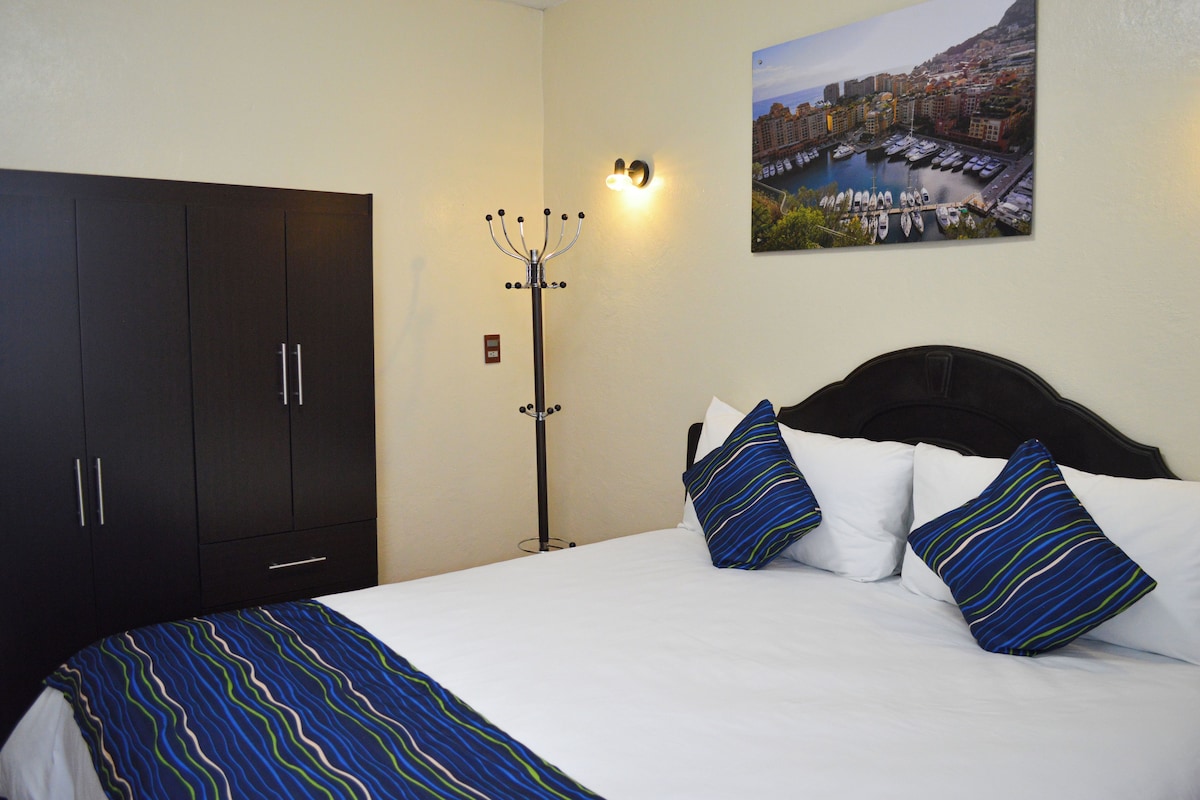 A hotel room with a bed, a dark wardrobe, a floor lamp, and a cityscape painting on the wall.