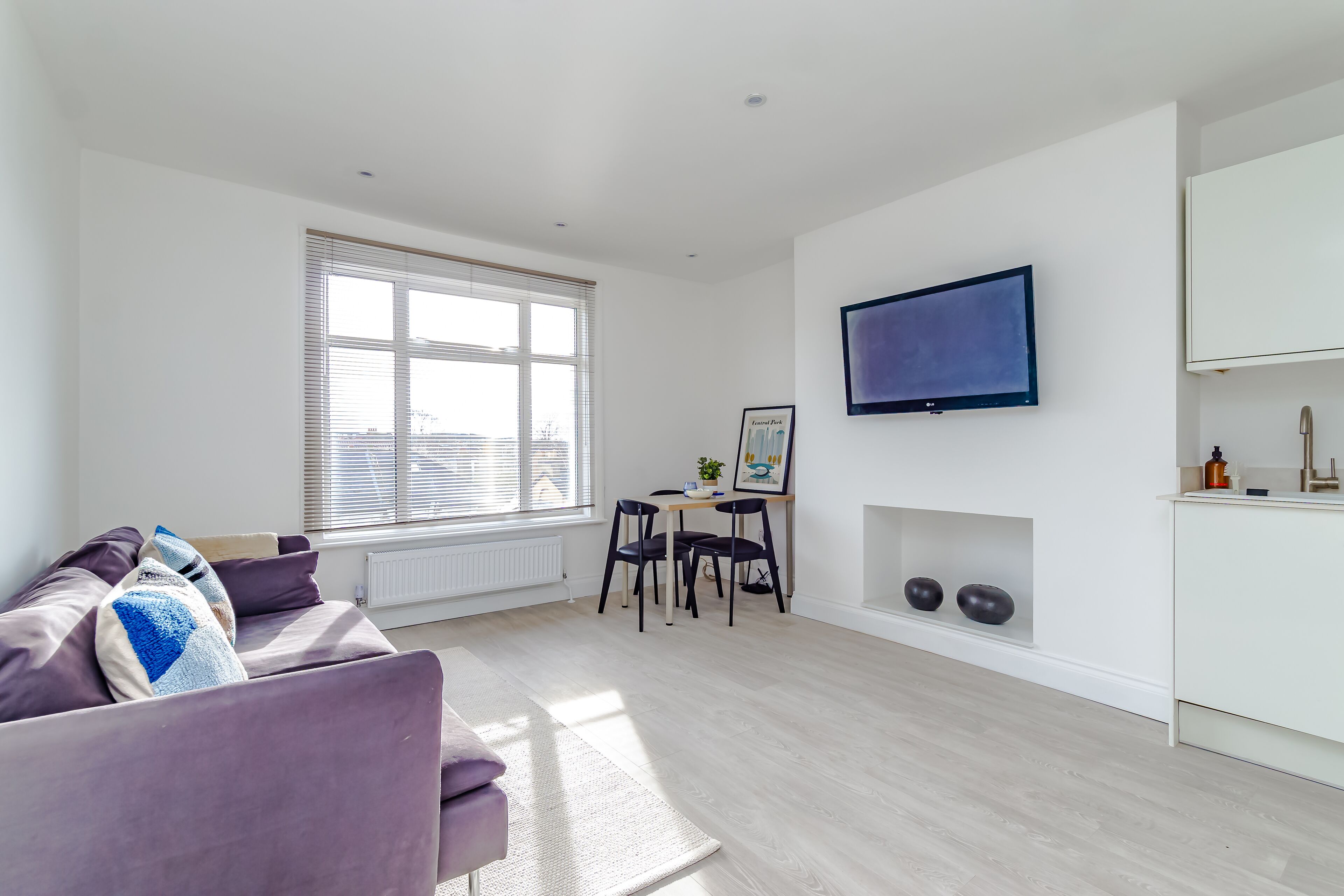 A modern living room with a sofa, a dining table with chairs, a TV mounted on the wall, and a kitchen area with cabinets.