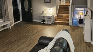 Una sala de estar con un sofá de cuero oscuro, una pequeña mesa blanca con una lámpara, y una escalera que lleva a un dormitorio.