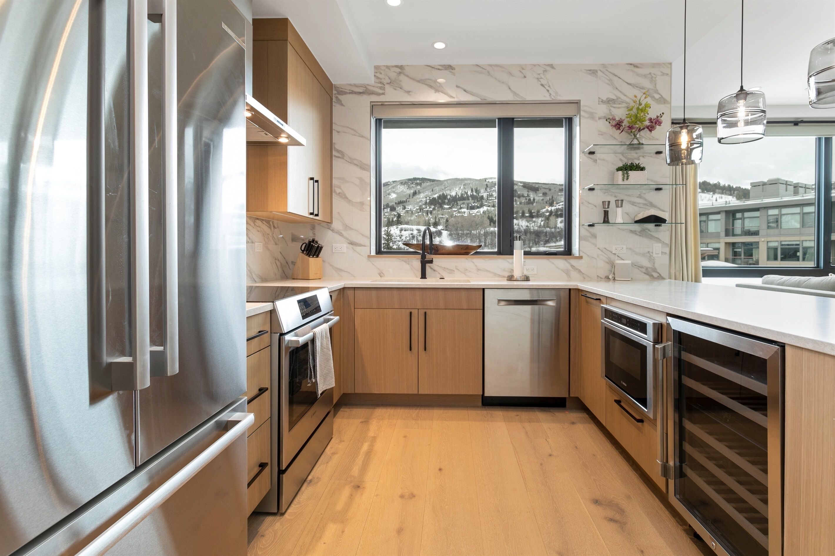 Une cuisine moderne dotée d’appareils électroménagers en acier inoxydable, de meubles en bois et d’une grande fenêtre offrant une vue sur des collines enneigées.