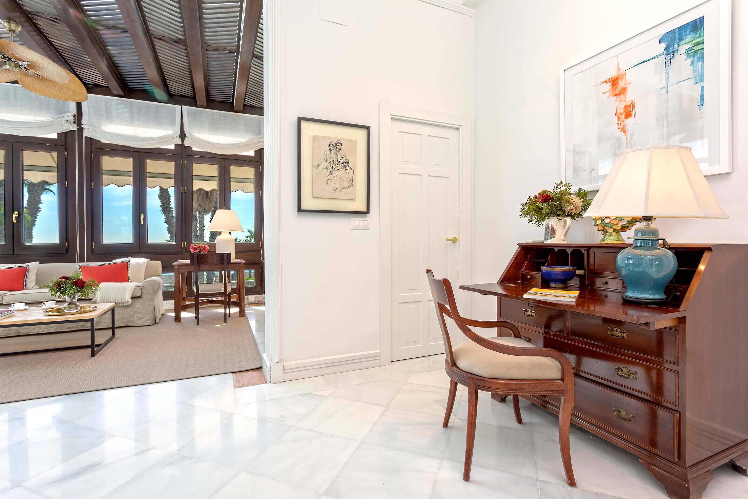 Amplia sala de estar con sofá, mesa de centro y un escritorio de madera con lámpara. Hay ventanas grandes y una puerta que da a otra habitación.