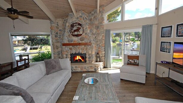 A cozy living room with a stone fireplace, a wooden ceiling fan, and a large window overlooking a garden. - Waterfront Home, Private Hot Tub/Boat Dock - Views! (South Lake Tahoe)
