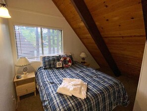 Une chambre avec un plafond en pente, un lit recouvert d’une couverture à carreaux, deux lampes de chevet, une table de chevet et une fenêtre avec des stores.
