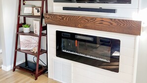 A living room with a flat-screen TV mounted above a white fireplace with a wooden mantel. A wooden ladder shelf with decorative items is placed against the wall. - Cute & cozy family-friendly Royal Oak ranch close to downtown and highways! (Royal Oak)