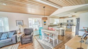 A spacious living area with a wooden ceiling, a dining table with chairs, a sofa with patterned cushions, and a kitchen with modern appliances.
