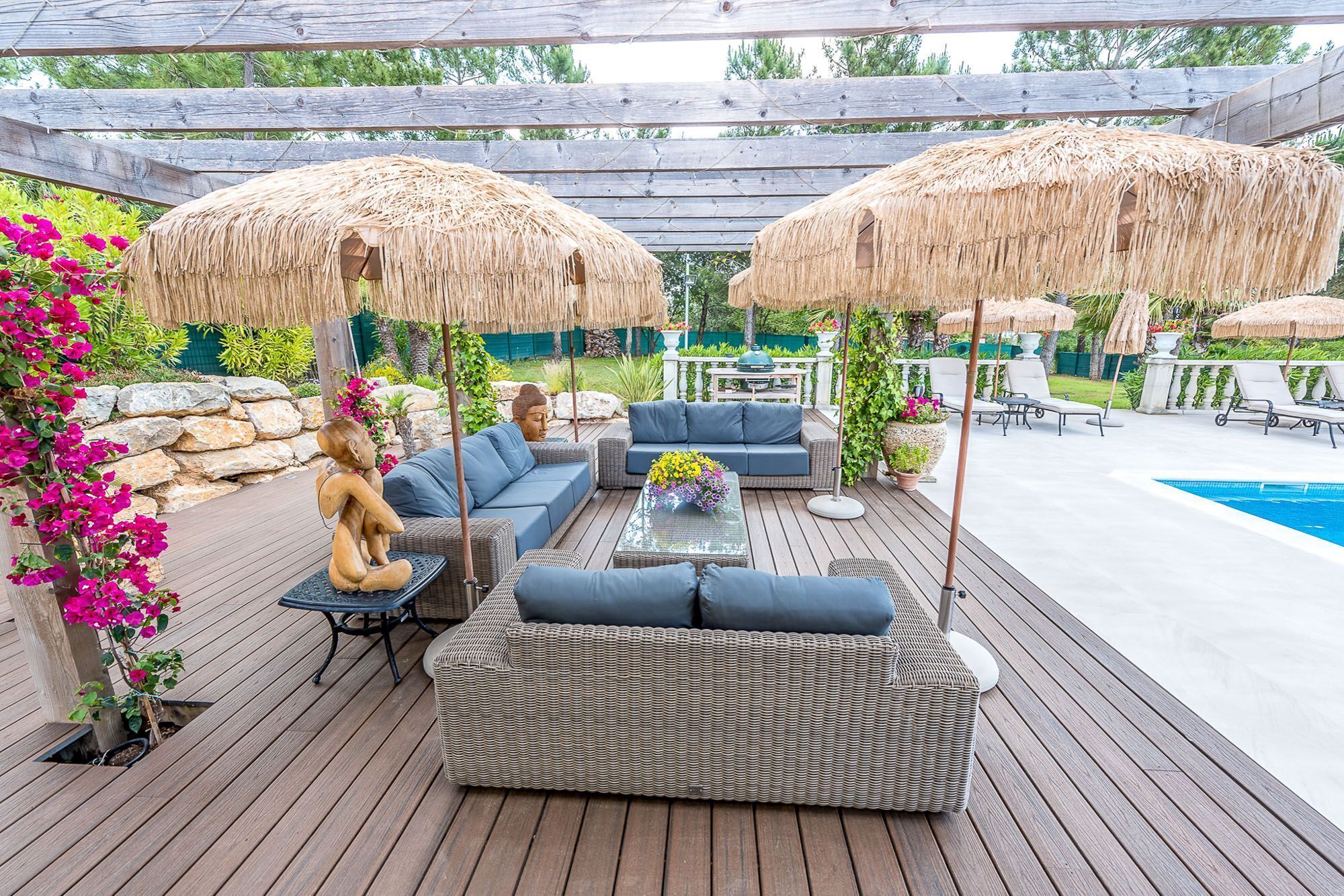 An outdoor deck with wicker furniture, a pool, and thatched umbrellas.