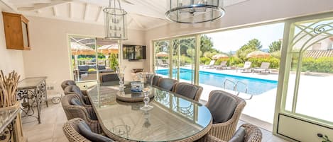 Un comedor soleado con una mesa de vidrio rodeada de sillas de mimbre, un ventilador de techo y vistas a una piscina.