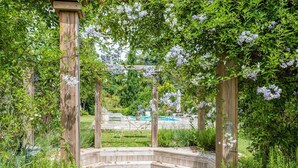 Una pérgola de madera con un brasero circular en el centro, rodeada de abundante vegetación y plantas en flor.