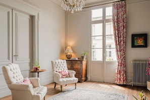 A room with two upholstered chairs, a wooden cabinet, a floor lamp, and a large window with patterned curtains.