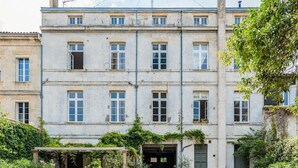 Un edificio de varios pisos con un jardín delantero, que incluye una pérgola con plantas en macetas.