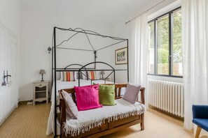 Een slaapkamer met een metalen bedframe, een houten bankje, een nachtkastje met een lamp en een raam met gordijnen.
