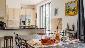 A modern kitchen with a central island, a dining area with a table set for two, and a view of the outdoors through large windows. - Mas Mayeaux - Six Bedroom Villa, Sleeps 12 (Vaison-la-Romaine)