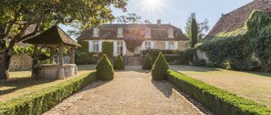 Un bâtiment traditionnel en pierre, doté d’une façade symétrique, d’un chemin de gravier menant à l’entrée principale et d’un jardin bien entretenu avec des haies taillées.
