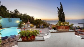 Un área de piscina en la azotea con sillones de descanso, plantas en macetas y vista al mar al atardecer.