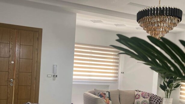 A living room with a beige sofa, a patterned throw, a chandelier, and a large window with blinds. - À 5 min du Parc Carthage Land! Magnifique Villa Avec Piscine Creusée à Louer (Hammamet)