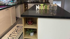 A modern kitchen with a black countertop, white cabinets, and a backsplash with patterned tiles. There is a small island with a 'NATURE' sign, a red pot, and a green plant on the shelf. - À 5 min du Parc Carthage Land! Magnifique Villa Avec Piscine Creusée à Louer (Hammamet)