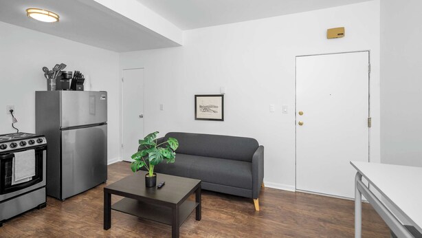 A compact kitchen with a refrigerator, stove, and dining table. A gray sofa and a small table with a plant are in the living area. - Northside Suites (Cincinnati)