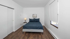 A bedroom with a bed, a nightstand, a lamp, a framed picture on the wall, and a window with blinds. - Northside Suites (Cincinnati)