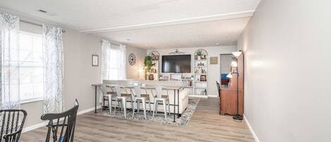 A dining area with a wooden table and chairs, a kitchen island with bar stools, and a TV.
