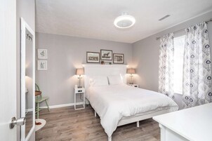Un dormitorio con cama, mesitas de noche, una silla, una ventana con cortinas estampadas y cuadros en la pared.