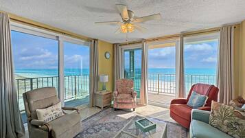 A spacious living room with a large window overlooking the beach, a ceiling fan, and a comfortable seating area with a coffee table.