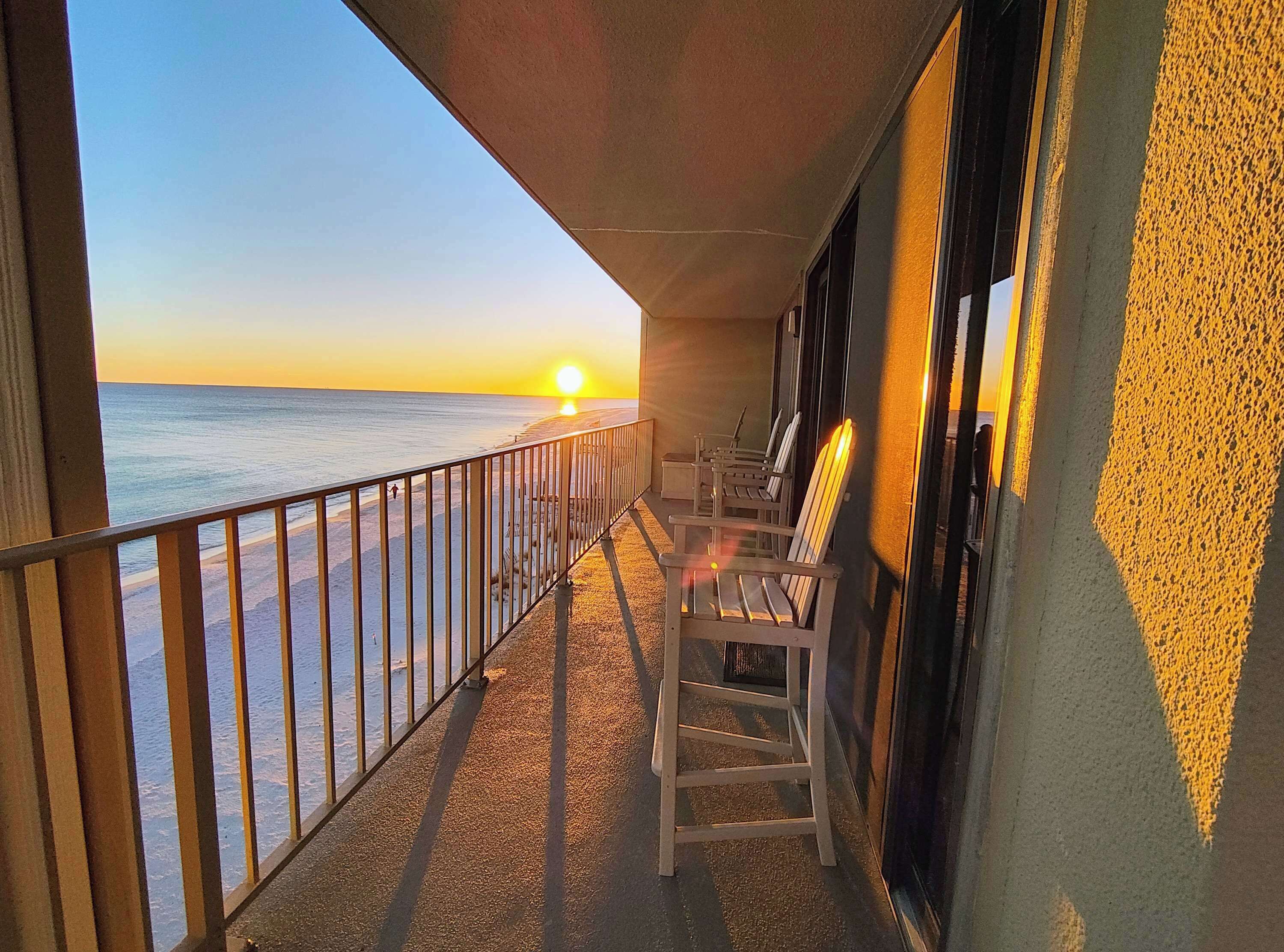 A balcony with a view of the sunset over the ocean, equipped with wooden chairs and a railing.