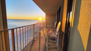 A balcony with a view of the sunset over the ocean, equipped with wooden chairs and a railing.