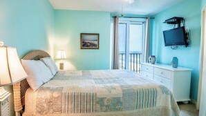 A bedroom with a bed, a nightstand, a lamp, a dresser, a TV, and a window with a view of the beach. - Oceanfront Oasis Beachfront Condo w/ Views! (Gulf Shores)