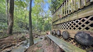 A wooden deck with a lattice railing, a bench, and a stream in a forested area.