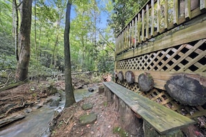 A wooden deck with a lattice railing, a bench, and a stream in a forested area. - Misty Creek Cabin Private 3 BDR w/ Game Room! (Blue Ridge)