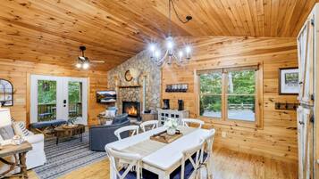 Interior de una cabaña de madera con comedor, asientos, chimenea y ventanas amplias.