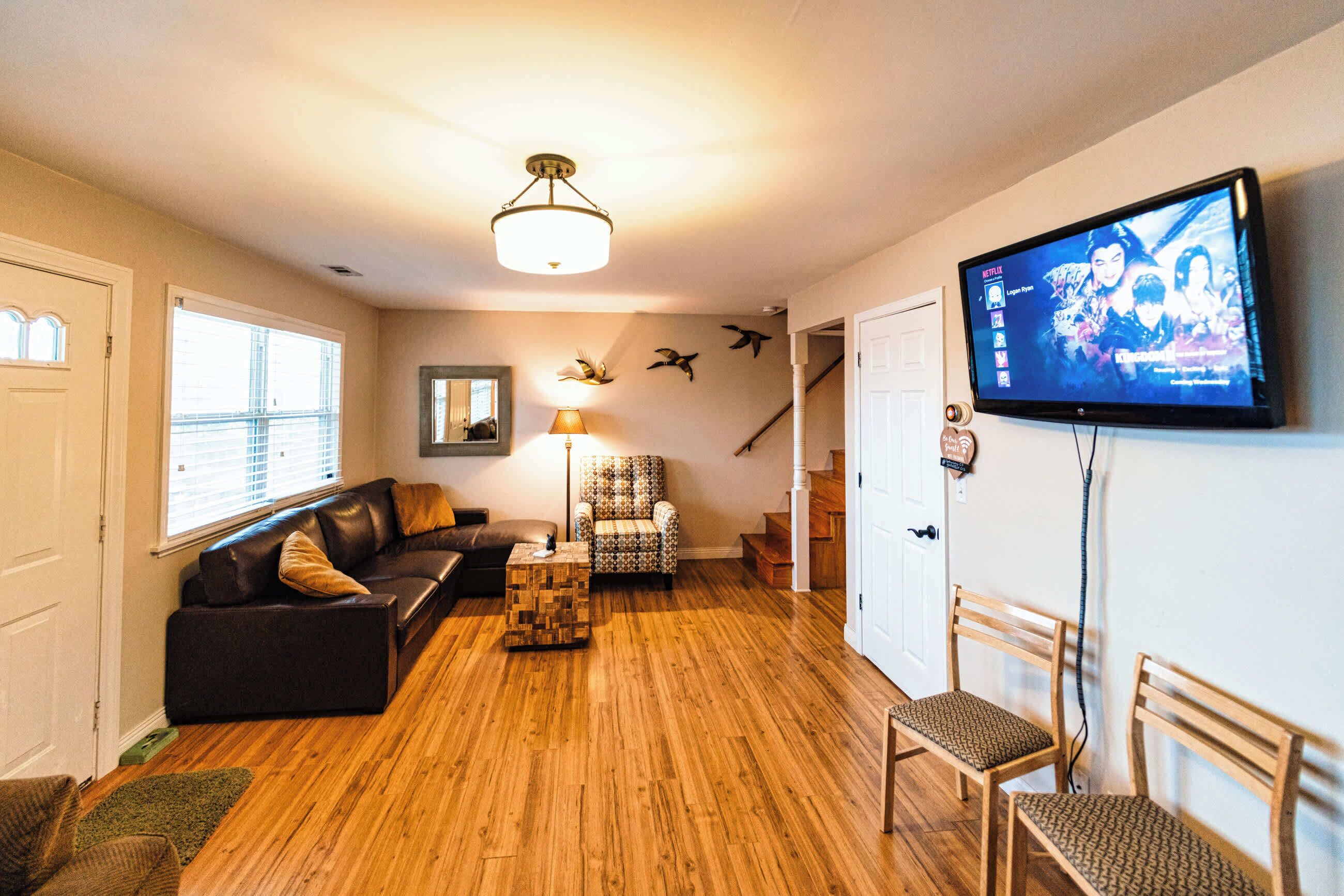 Una sala de estar con piso de madera, un sofá de cuero negro, un sillón estampado, una mesa de centro de madera y un televisor montado en la pared.