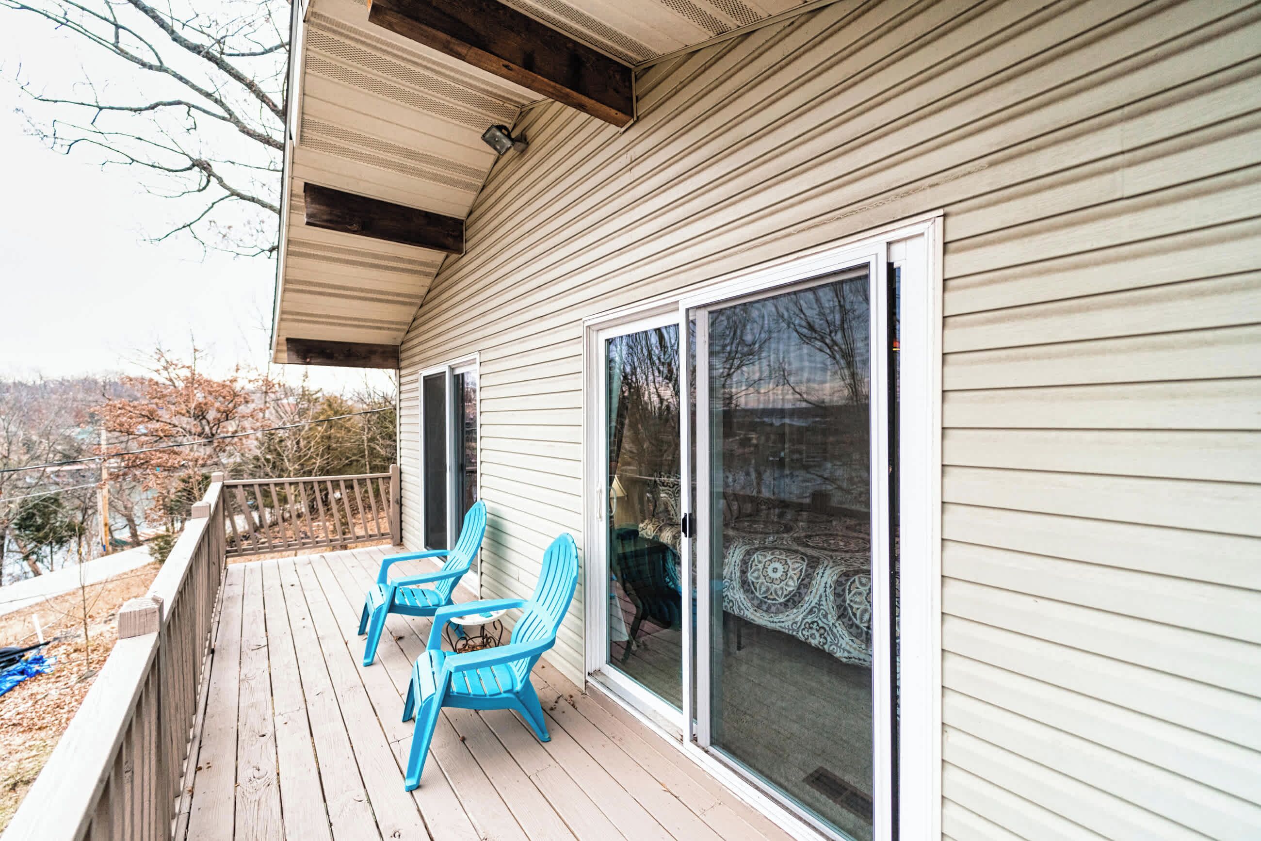 A wooden deck with two blue Adirondack chairs, a sliding glass door, and a view of trees and hills.