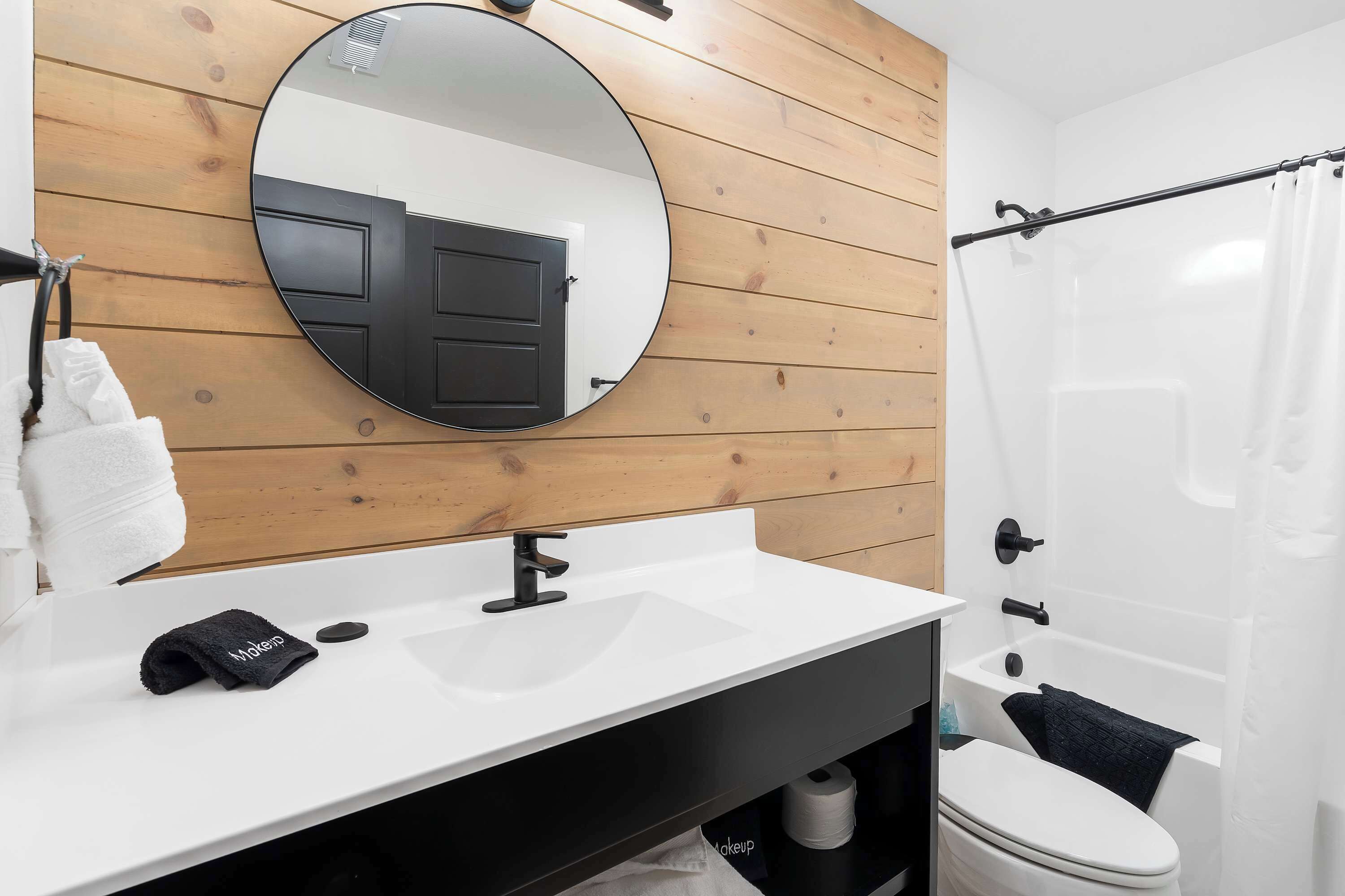 Une salle de bain moderne avec un mur recouvert de panneaux en bois, un miroir rond, un lavabo blanc, une serviette noire et des toilettes blanches.