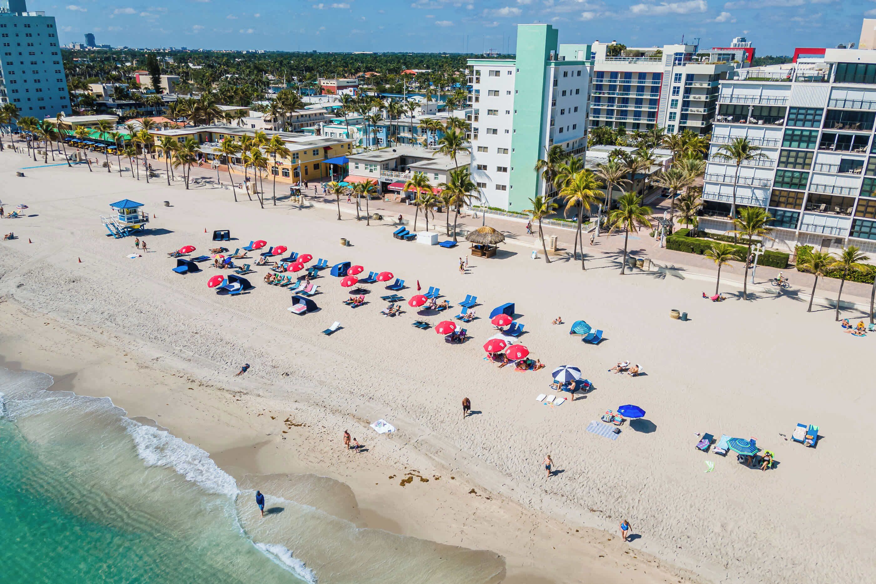 Una playa con numerosos paraguas y tumbonas, palmeras y edificios al fondo.