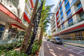 A modern hotel corridor with palm trees, a coffee shop, and a van parked on the side. - Condo Close to the Beach With Rooftop Pool & Gym (Hollywood)