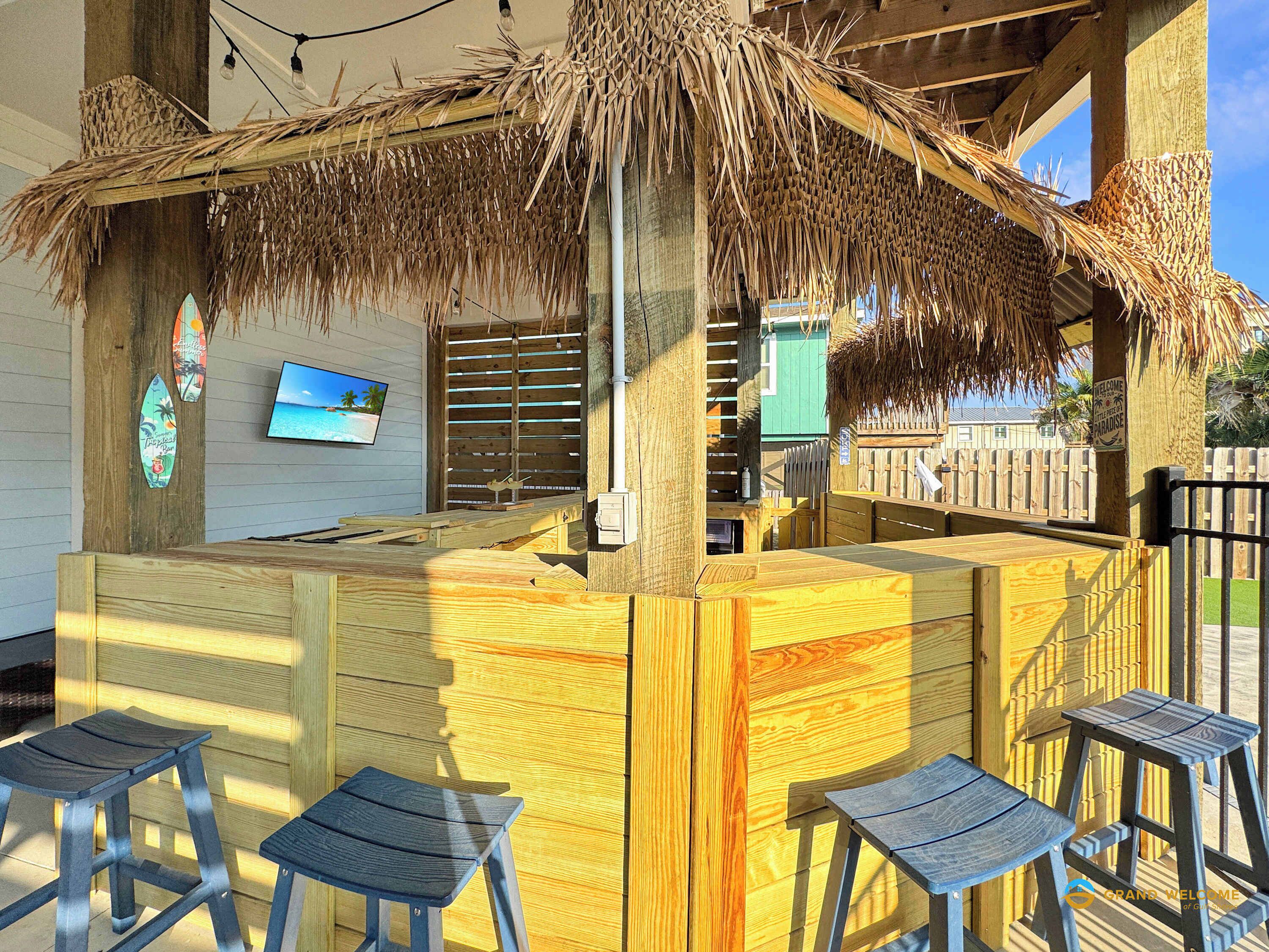 Un área de bar con barra de madera, techo de paja, taburetes de madera y una pantalla de televisión en la pared.