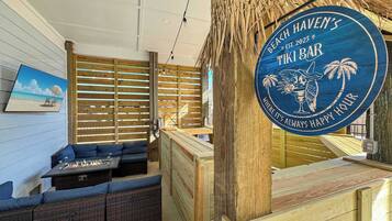 Un bar tiki con interior de madera, un letrero que dice "Beach Haven's Tiki Bar" y vistas de la playa a través de una ventana.